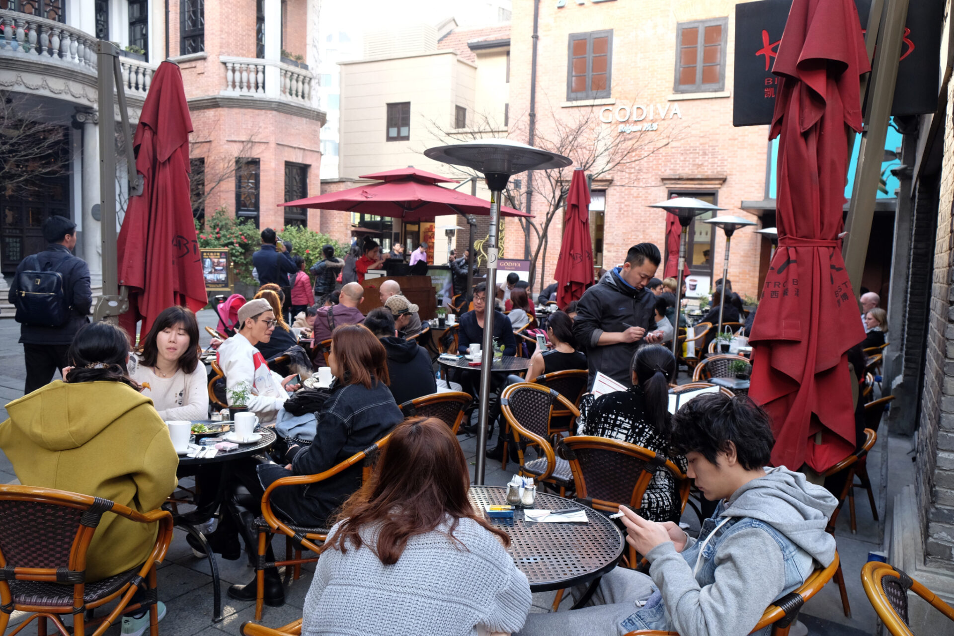 Außengastronomie im historischen Stadtviertel Xintiandi in Shanghai mit Tischen, roten Sonnenschirmen und zahlreichen Gästen.